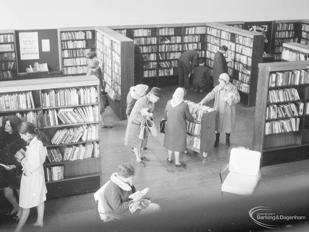 London Borough of Havering Central Library, Romford, showing part of the lending department, with bookcases, library users, and women looking at books on trolley, 1967