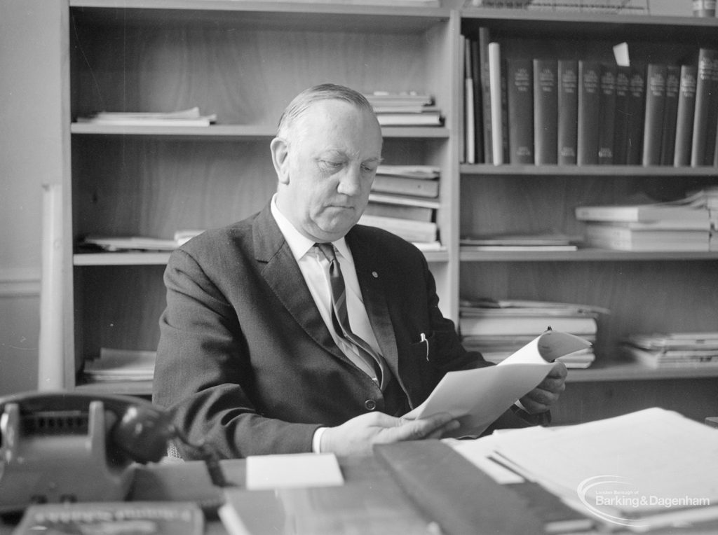 London Borough of Barking Borough Librarian Mr E W McManus FLA, at his desk and reading an open booklet, 1972