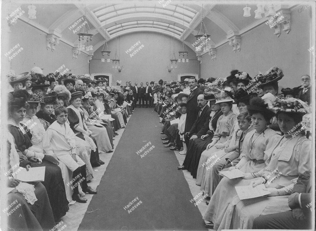 The official opening of Hackney Central Library by their Royal Highnesses the Prince and Princess of Wales. Shows a crowd of adults and children sat in a long room on the 1st floor of the building in smart Edwardian dress,1908