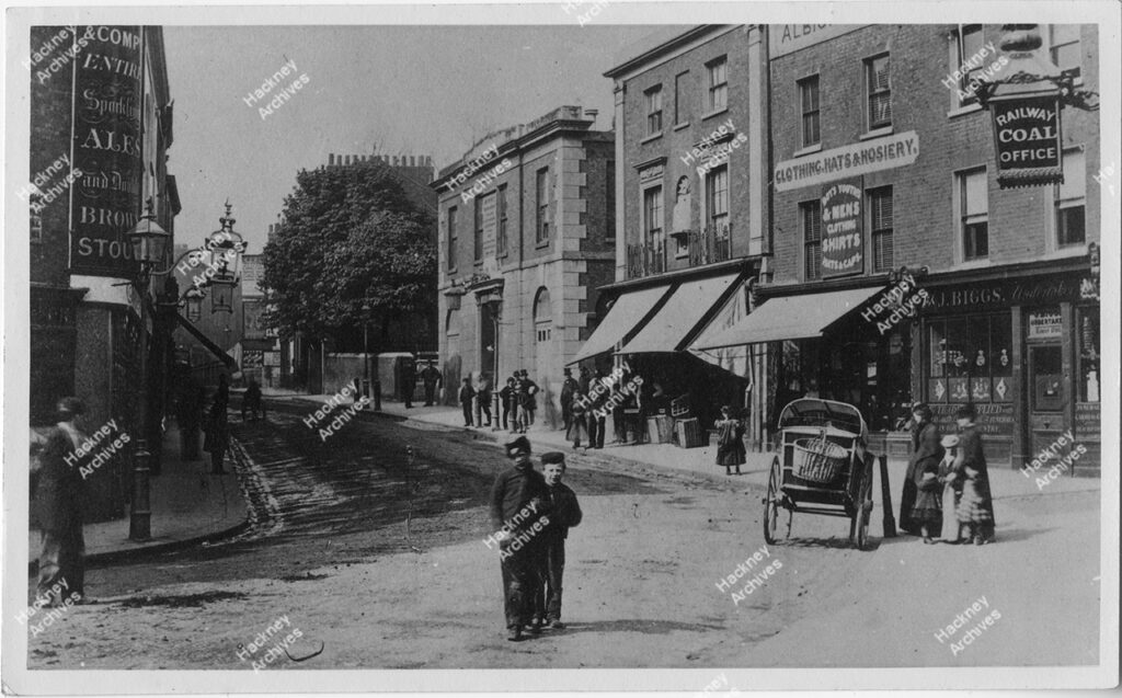 Mare Street, Hackney looking north from railway bridge. Includes Eight Bells public house, J. Biggs undertakers and old town hall. Children, men and woman in Victorian dress and a hand cart, c.1880
