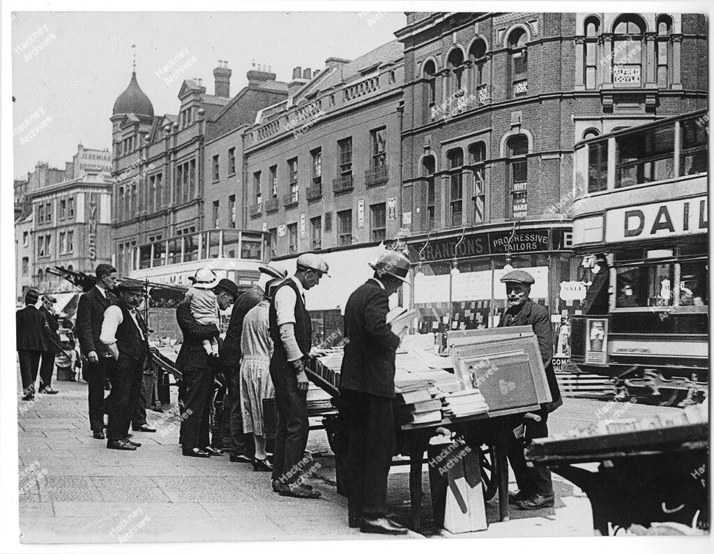 Bookstalls and sellers on Shoreditch High Street looking north east. Includes Jeremiah Rotherham’s factory, Brandons Progressive Tailors, c.1930.
