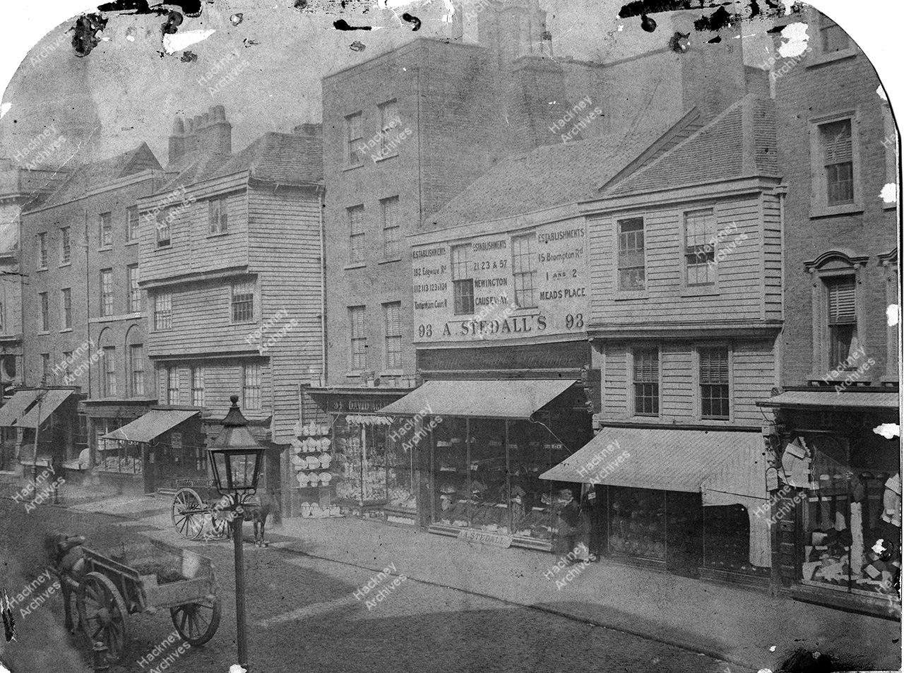 East side of High Street, Shoreditch nearby the Church. Horse drawn carts outside shops. 1875.
