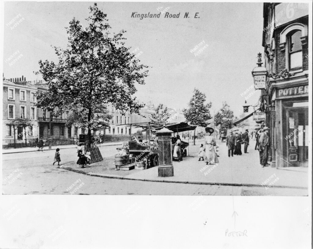 Corner of Kingsland Road and Richmond Road, Hackney, c.1905. Includes number 442 The Potter Press printing works (the original Gazette Office). Mr Potter in grey top hat in foreground. Market stalls and horse drawn cart