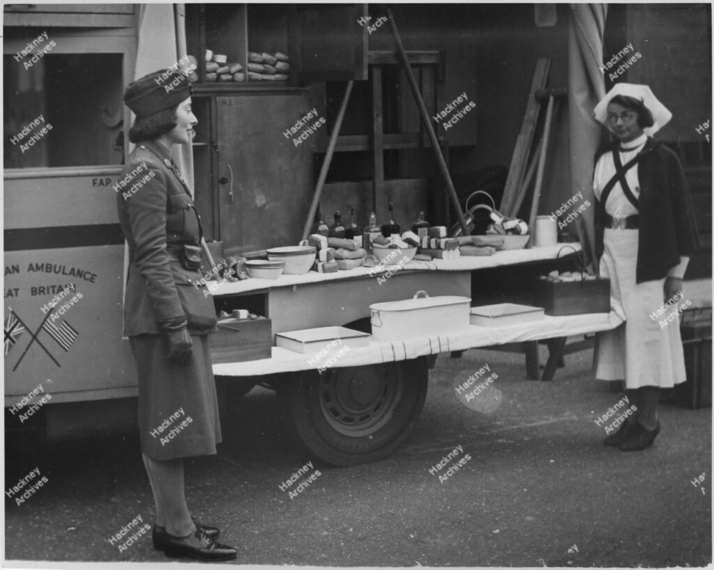 WW2 War Weapons Week exhibition stand, Stoke Newington Town Hall, Stoke Newington, Hackney. 1941. Includes a nurse and a member of the Women’s Auxiliary Army Corp (WAAC) in front of a ambulance and a display of medical supplies.