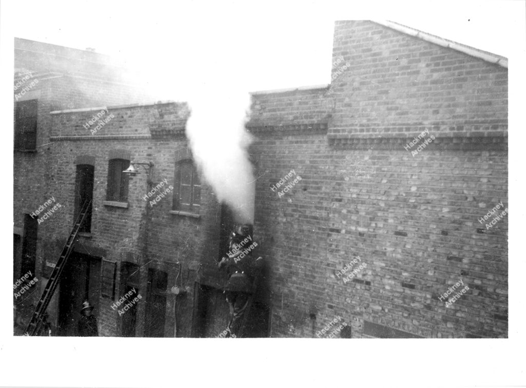 Fire in premises backing on to Batley Place (52-58 Stoke Newington High Street).Firefighters on ladder entering window with smoke coming from it. Hackney, c.1960.