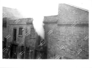 Fire in premises backing on to Batley Place (52-58 Stoke Newington High Street).Firefighters on ladder entering window with smoke coming from it. Hackney, c.1960.