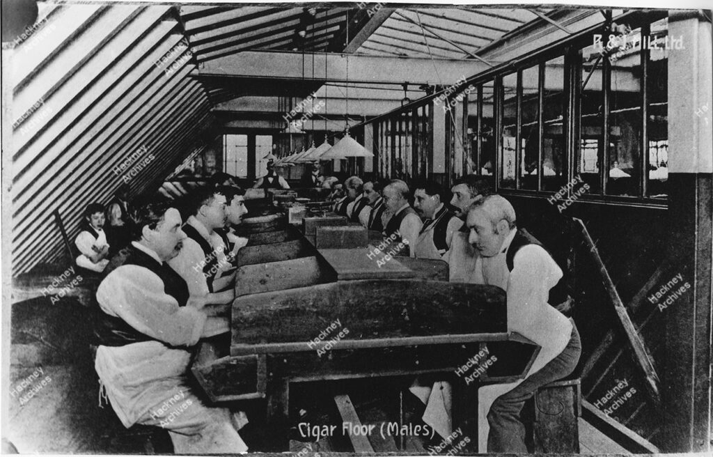 Workers at R & J Hill Ltd, Tobacco manufacturers. Men seated at a row of work tables on the ‘Cigar Floor’. Boys are seated behind the men. c.1850.