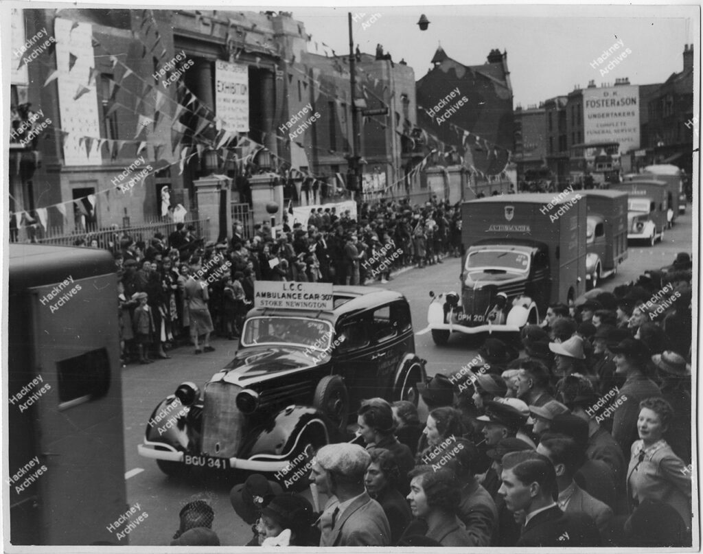 WW2 War Weapons Week exhibition stand, Stoke Newington Town Hall, Stoke Newington, Hackney. 1941. Exhibition stands at Stoke Newington town hall (including ‘Help Stoke Newington to buy bombs for Berlin’), procession in Church Street, ambulance display, Mayor and civic group in town hall front area. Old St Mary’s Church at rear and members of Stoke Newington ARP (Air Raid Patrol)